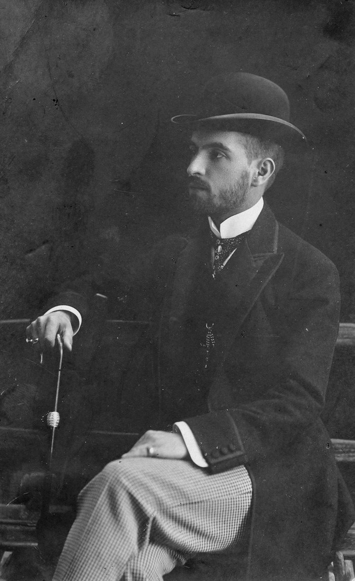 Photographie de studio d’un jeune homme barbu en chapeau melon et manteau sombre, assis de profil et appuyé sur une canne.