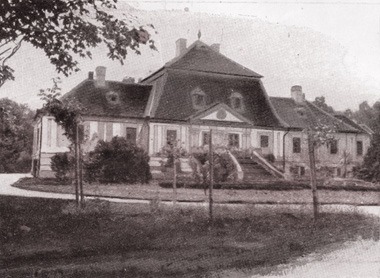 Photographie en noir et blanc du manoir de Rudna : une maison à deux étages sous un toit mansardé, avec un avant-corps central à fronton, un escalier à double volée et des arbres devant.