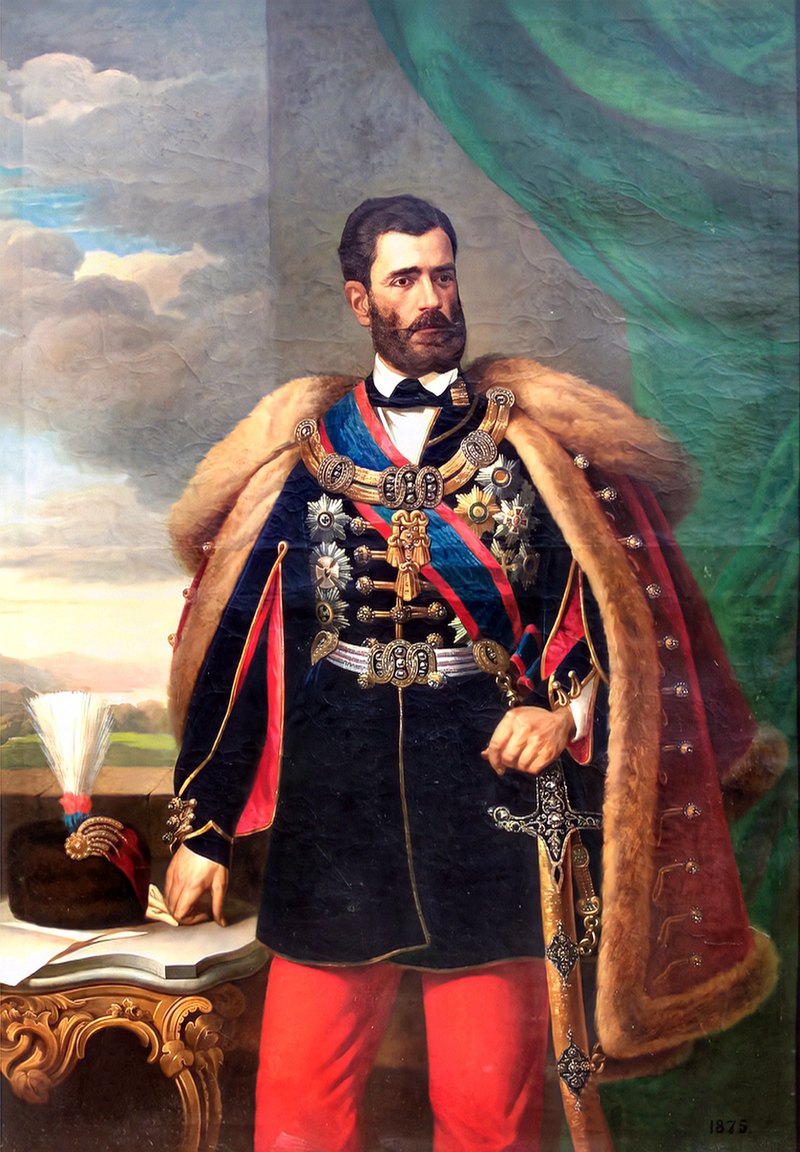 Portret u punoj figuri kneza Mihaila Obrenovića u tamnoj husarskoj tunici sa odlikovanjima i ogrtačem oivičenim krznom, sa kapom sa perjanicom na stolu pored njega; u uglu je upisana godina 1875.