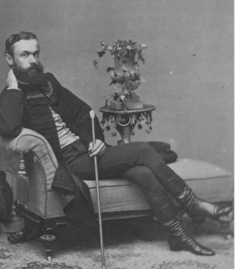 Photographie de studio d’un jeune homme barbu en veste courte sombre et bottes, allongé sur une chaise longue avec une canne, un vase de fleurs sur une table derrière lui.