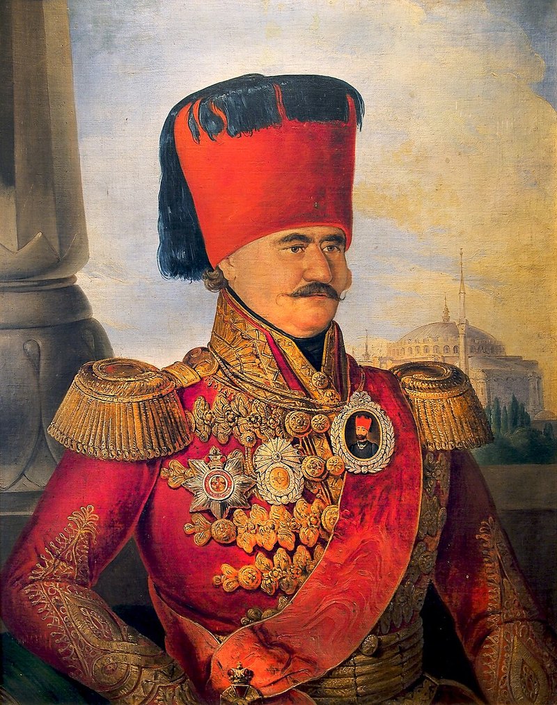 Slika kneza Miloša Obrenovića u visokom crvenom fesu sa crnom kićankom, u crvenoj uniformi sa zlatnim epoletama i odlikovanjima, pred kamenim stubom i džamijom sa kupolom.