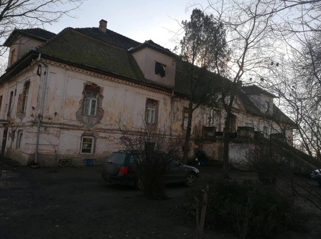 Vue latérale du manoir : une maison à deux niveaux sous de hauts toits de tuiles, avec une lucarne et un crépi usé, une voiture stationnée devant.