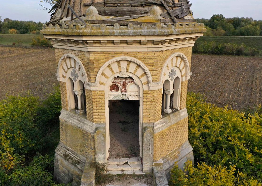 Vue aérienne d’une tour funéraire octogonale en brique jaune, aux fenêtres en plein cintre et au dôme en bois endommagé, entre des buissons et des champs labourés.