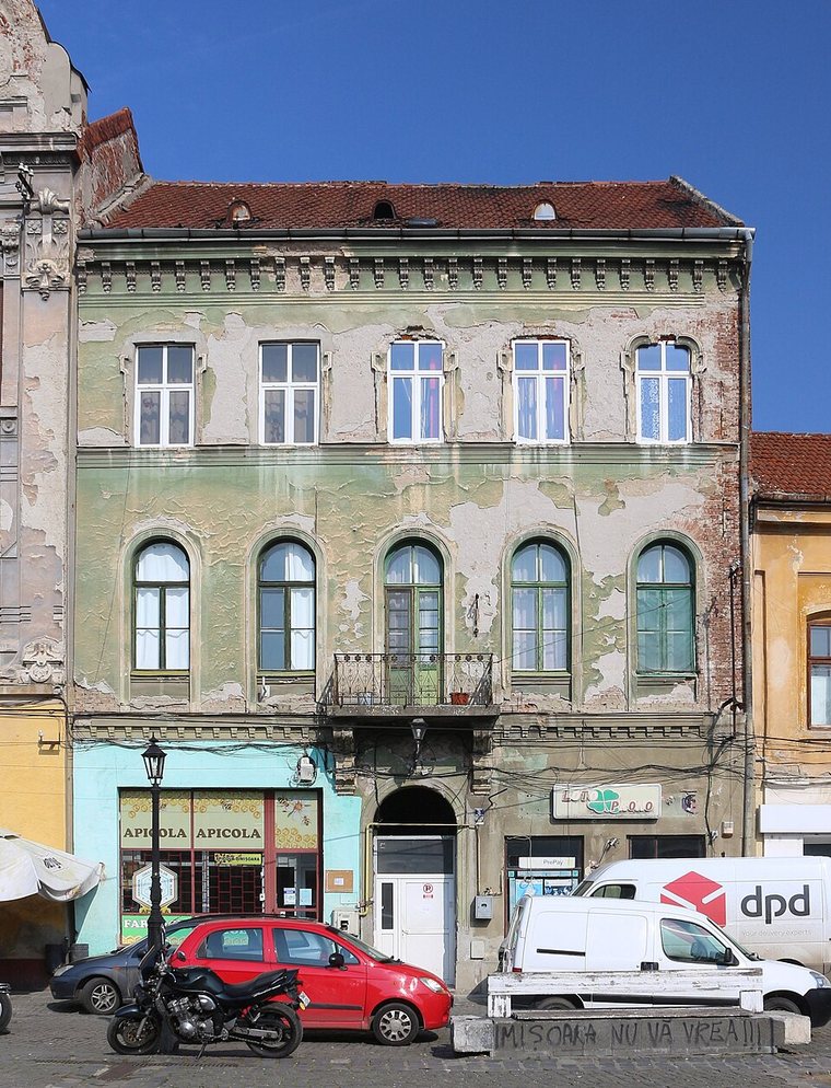 Façade d’un hôtel particulier du XIXe siècle à trois niveaux, sur la Piața Traian à Timișoara, avec des fenêtres cintrées au rez-de-chaussée, un petit balcon en fer et un crépi vert-gris qui s’écaille.